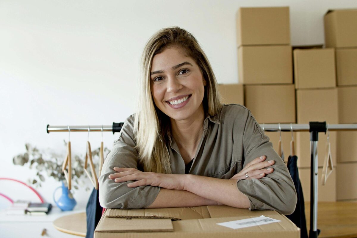 Frau will selbstgemachtes verkaufen, lehnt sich auf einem Karton, im Hintergrund Kartons mit Handarbeit f&uuml;r den Verkauf im Onlineshop